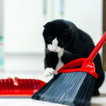 Black-and-white cat pawing at the red O-Cedar broom head next to a red dustpan on a bright kitchen floor