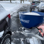 Hand-held blue Chapin ice-melt spreader filled with white granules in a snowy driveway next to parked cars.