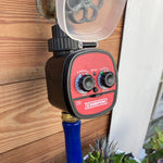 Chapin water timer mounted on an outdoor faucet with protective cover, connected to a hose fitting against a wooden wall.