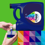 Hands pouring dark blue cheer colorguard detergent from a large jug into a measuring cap over a colorful patterned surface