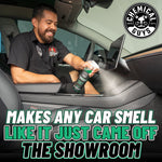 Person spraying a Chemical Guys New Car Smell bottle inside a car interior with large brand logo and bold tagline