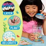 Child assembling a terrarium, holding the decorated lid while jar with capybara figurines, stickers and kit materials sit on table.