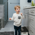 Toddler standing in a bathroom brushing teeth with a gray otter-shaped toothbrush