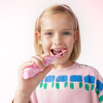 Young girl using a pink battery toothbrush in a lifestyle shot, smiling while brushing against a pale pink background.