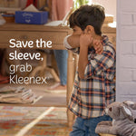Child coughing into an elbow in a home setting with a Kleenex box visible on a table in the background