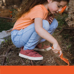 Child crouching and using an orange toy cultivator in soil while wearing casual clothes, focused on hands and tool action.