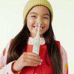 Smiling child holding a green Ordo battery toothbrush vertically toward the camera, wearing a yellow knit hat and red outfit.
