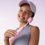 Smiling child holding a pink Ordo sonic toothbrush with a cow-themed Squishmallows brush cap against a pale background.