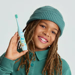Child model holding a mint green Ordo kids electric toothbrush decorated with Squishmallows characters, smiling against a pale background