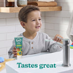Young child holding a tube of Tom’s Kid’s Natural toothpaste at a bathroom sink, smiling while reaching for a toothbrush; overlay text reads Tastes great.