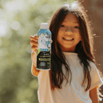 Smiling child holding a can of Trek kids insect repellent toward the camera outdoors with soft bokeh background