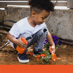 Young child kneeling outdoors holding an orange toy spray bottle in one hand and an orange trowel in the other near flowers.