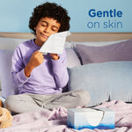 Child seated on a bed using a tissue with an open Puffs tissue box nearby and text reading ‘Gentle on skin’