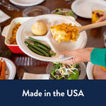 Hand holding a Chinet three-compartment disposable plate with green beans, roll and a scoop of casserole at a dining table.