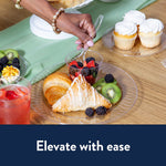 Hand picking up a clear plastic fork from a cup of berries on a plate with croissant, kiwi and cupcakes nearby
