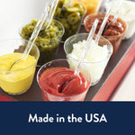 Clear plastic spoons and forks placed in small condiment cups holding ketchup, mustard, onions and other toppings