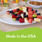 White Chinet plate serving fruit skewers on a table, with blurred snack cups in the background and 'Made in the USA' banner.