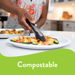 Lifestyle photo of a person using tongs to place food on a white Chinet platter on a kitchen counter, 'Compostable' text area