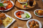 Tabletop party spread showing white oval disposable platters filled with sliders, wings, chips and dipping cups