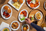 Overhead view of a table with white oval disposable platters of wings, sliders and pretzels; hand reaching for food
