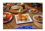 Tabletop party scene with Chinet white square plates holding sliders, chips and assorted appetizers on a wood table