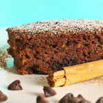 Close-up of a spiced chocolate chip loaf slice dusted with powdered sugar, with a cinnamon stick and chocolate chips in foreground.