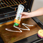 Scrub Daddy Cif cream being squeezed onto an oven door surface with a yellow-and-pink sponge nearby