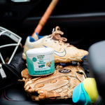 Citrus Magic Sea Breeze solid air freshener tub placed on a leather car seat next to a baseball glove and cleat