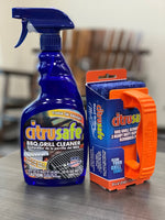 Citrusafe blue spray bottle next to an orange-handled grill scrubber on a tabletop with blurred background.