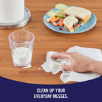 Hand wiping spilled milk on a wood table with a Sparkle paper towel beside a glass and sandwich plate