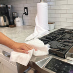 Hand cleaning stainless steel stovetop edge with a white Tear N Clean towel and roll visible on counter