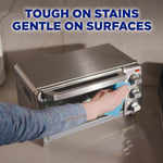 Person wiping a stainless steel toaster oven door with a blue cloth and text 'Tough on stains gentle on surfaces' above