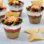 Clear plastic cups filled with layered dip and topped with olives, cheese, and a tortilla chip garnish