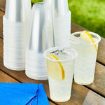 Outdoor tabletop scene showing clear disposable cups filled with lemonade and stacked unfilled cup stacks behind