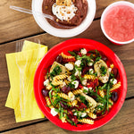 Top-down lifestyle scene with clear plastic fork next to a red bowl of pasta and dessert cups on a wooden table