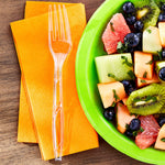 Clear disposable plastic fork placed on orange napkins beside a green plate of mixed fruit in a top-down view