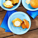 Styled overhead scene showing clear plastic knives on blue paper plates with muffins on a wood surface
