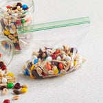 Clear sandwich bag with mixed nuts and candy pieces inside next to glass jars, showing green double zipper closure