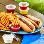Lifestyle photo of clear disposable sauce cups with lids beside fries and hot dogs on a picnic table