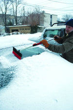 Person clearing snow from a car windshield with a telescoping broom showing red bristles and foam grip