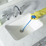 Gloved hand holding a blue Clorox drain stick down toward a white sink drain, demonstrating product use on a marble countertop.