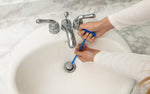 Hands using a blue Clorox drain-cleaning stick to fish debris from a white bathroom sink drain in a lifestyle scene.