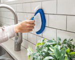Person cleaning kitchen tile backsplash with Clorox handheld diamond scrubber near a sink and faucet