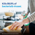 Close-up lifestyle image of hands wiping a stovetop with a yellow cloth while a blurred Clorox spray bottle is used in background and claim about killing 99.9%.