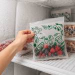 Close-up of a hand holding a Lunchskins compostable freezer bag filled with berries inside a frosty freezer shelf with other containers nearby.