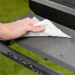 Close-up of a hand wiping small crumbs and debris from a gray metal grill shelf using a white cleaning cloth.
