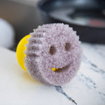 Close-up of a gray smiley-face scrub pad attached to a yellow Scrub Hub resting on a countertop