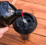 Close-up of dark jug pouring clear torch fuel into a black torch head wick assembly on a wooden surface