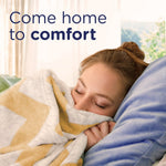 Woman cuddling a blanket on a couch with text 'Come home to comfort' suggesting a cozy lifestyle scene.