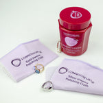 Connoisseurs jewelry cleaner jar with two polishing cloths and assorted rings arranged on a white surface